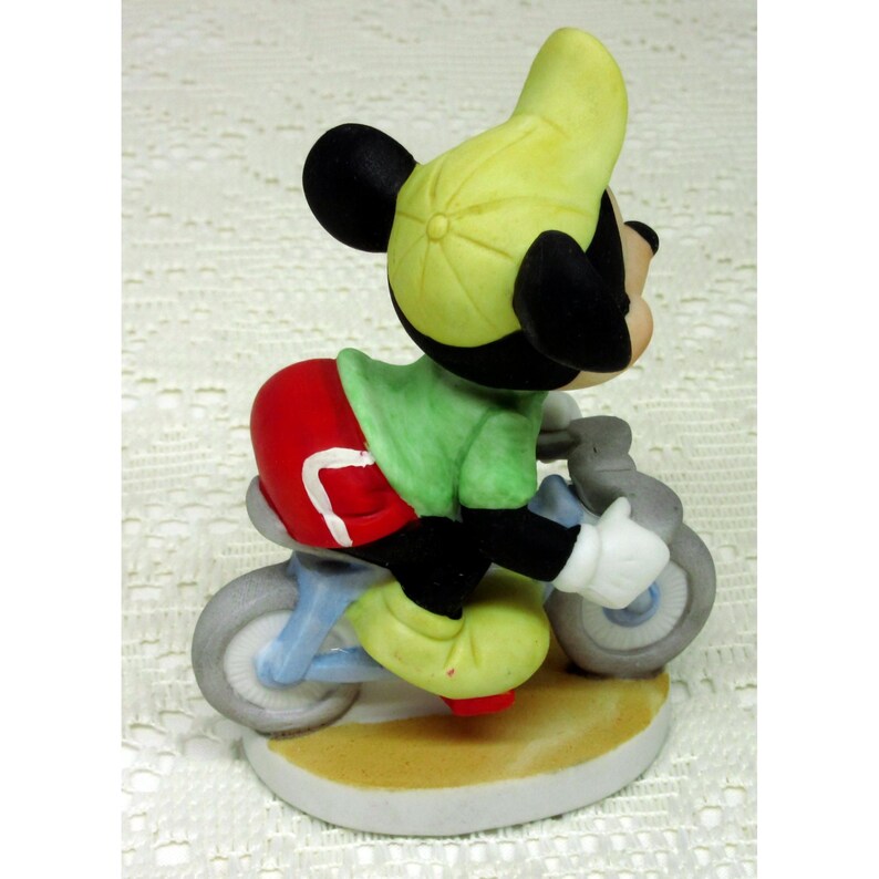 Puede incluir: Figura de cer&aacute;mica de Mickey Mouse en bicicleta. Mickey viste camisa verde, pantal&oacute;n corto rojo y gorra amarilla. La bicicleta es azul claro con ruedas grises. La figura est&aacute; sobre una base blanca con borde beige.