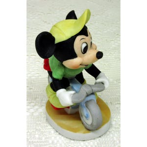 Puede incluir: Una figura de cer&aacute;mica de Mickey Mouse montando en bicicleta azul claro y gris. Mickey lleva una camisa verde, un sombrero amarillo, pantalones cortos rojos y guantes blancos. La figura est&aacute; sobre una base ovalada beige y blanca.
