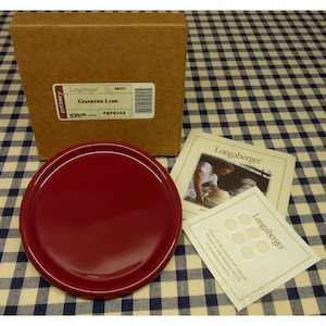May include: A round, deep red ceramic coaster lid by Longaberger. It is accompanied by a brown cardboard box labeled Coaster Lids Paprika, a small informational booklet with a photograph, and a sheet of six small, circular adhesive felt pads.