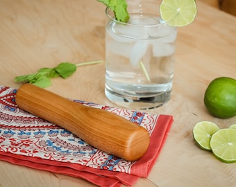 Puede incluir: Un mortero de madera descansa sobre una servilleta estampada en rojo y blanco junto a un vaso de agua con hielo, menta y una rodaja de lima. También hay una lima entera y rodajas de lima. El texto "EVANS WOODSHOP" y "EVANSWOODSHOP.COM" es visible.