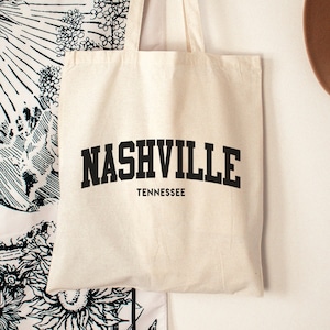 May include: A white canvas tote bag with black text that reads "NASHVILLE TENNESSEE". The bag is hanging on a white wall with a black and white tapestry and a brown hat in the background.