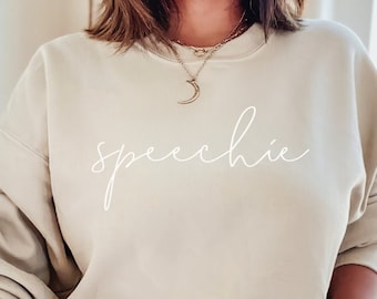 Puede incluir: Una sudadera beige claro con la palabra "speechie" escrita en cursiva blanca en la parte delantera. La sudadera la lleva puesta una persona con un collar de media luna.