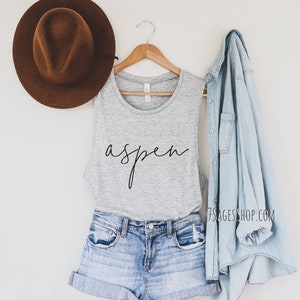 May include: A gray tank top with the word "aspen" written in black script. The tank top is paired with denim shorts and a light blue denim jacket. A brown fedora hat is on the left side of the image.