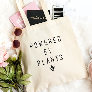 May include: A beige tote bag with the text "POWERED BY PLANTS" printed in black. The bag is filled with various items, including a pink notebook, a black notebook, a white phone, and a pair of sunglasses.