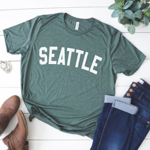 May include: A green t-shirt with the word "SEATTLE" printed in white on the front. The shirt is laid out on a white wooden surface with a pair of brown boots and blue jeans.