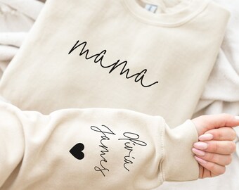 Puede incluir: Una sudadera beige con la palabra "mama" impresa en la parte delantera y "Olivia-in-James" con un corazón negro impreso en la manga.