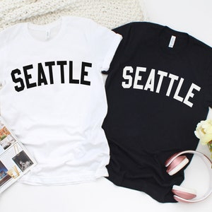 May include: Two t-shirts, one white and one black, with the word "SEATTLE" printed in a bold, arched font. The white shirt has black text, and the black shirt has white text.
