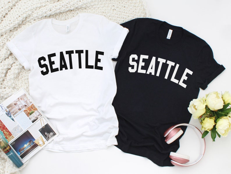 May include: Two t-shirts, one white and one black, with the word "SEATTLE" printed in a bold, arched font. The white shirt has black text, and the black shirt has white text.