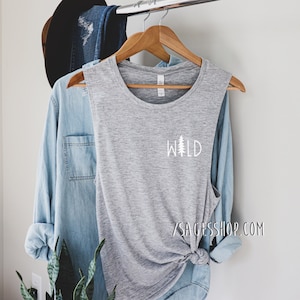 K&ouml;nnte beinhalten: Graues &auml;rmelloses Muscle-Tanktop mit einer wei&szlig;en Grafik eines Kiefernbaums und dem Wort "WILD" auf der Vorderseite. Das Tanktop h&auml;ngt an einem Holzb&uuml;gel.