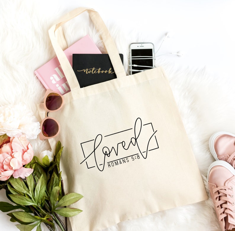 K&ouml;nnte beinhalten: Eine beige Canvas-Tragetasche mit schwarzen Henkeln und dem Text "Loved" in schwarzer Schrift mit einem schwarzen Rechteck darum. Der Text "R&ouml;mer 5:8" ist in Schwarz unter dem Rechteck geschrieben. Die Tasche ist gef&uuml;llt mit einem rosa Notizbuch, einem schwarzen Notizbuch mit dem Wort "Notebook" darauf, einem wei&szlig;en Telefon und einer Sonnenbrille. Die Tasche liegt auf einer wei&szlig;en, flauschigen Oberfl&auml;che.