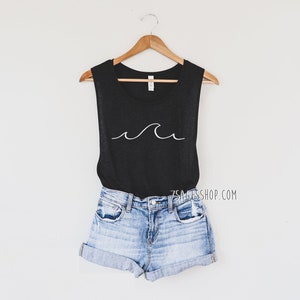 May include: A black sleeveless tank top with a white wave graphic. The tank top is hanging on a wooden hanger and is styled with denim shorts.