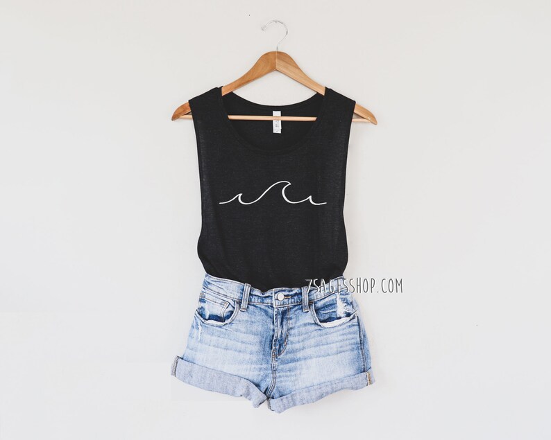 May include: A black sleeveless tank top with a white wave graphic. The tank top is hanging on a wooden hanger and is styled with denim shorts.