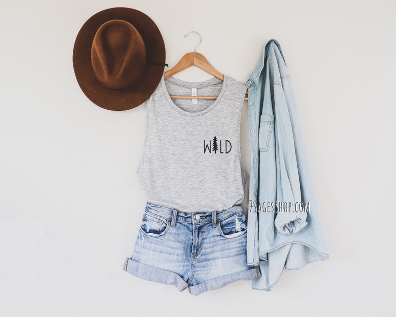 K&ouml;nnte beinhalten: Ein graues Tanktop mit dem Wort "WILD" und einer kleinen Tannenbaumgrafik auf der Vorderseite. Das Tanktop wird mit Jeans-Shorts und einer hellblauen Jeansjacke kombiniert.