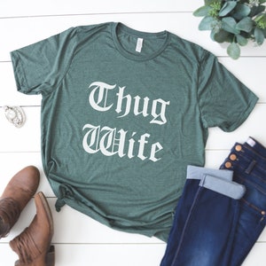 May include: A green t-shirt with the text "Thug Wife" in white lettering. The shirt is laid flat on a white wooden surface with a pair of brown boots and blue jeans.