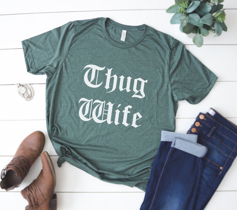 May include: A green t-shirt with the text "Thug Wife" in white lettering. The shirt is laid flat on a white wooden surface with a pair of brown boots and blue jeans.