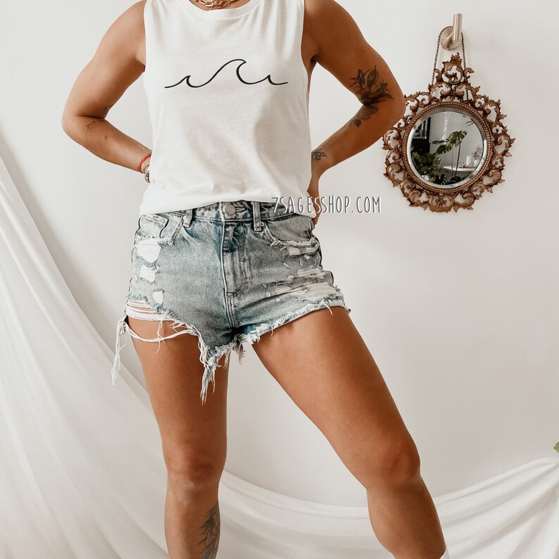 May include: A white sleeveless shirt with a simple black wave design. The shirt is paired with light blue denim shorts with distressed details.