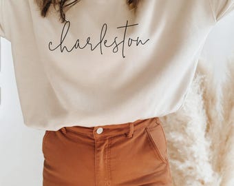 May include: A beige sweatshirt with the word "Charleston" in a cursive font. The person is wearing brown pants. The background includes a decorative plant.
