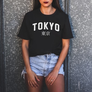 K&ouml;nnte beinhalten: Ein schwarzes T-Shirt mit wei&szlig;em Text, auf dem "TOKYO" und "東京" in japanischen Schriftzeichen steht.