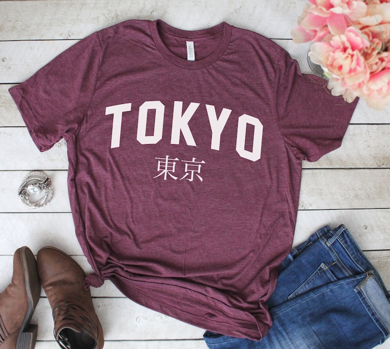 K&ouml;nnte beinhalten: Ein maroonfarbenes T-Shirt mit dem Wort "TOKYO" in wei&szlig;en Blockbuchstaben und den japanischen Schriftzeichen f&uuml;r "Tokyo" darunter. Das Shirt liegt auf einer wei&szlig;en Holzoberfl&auml;che.