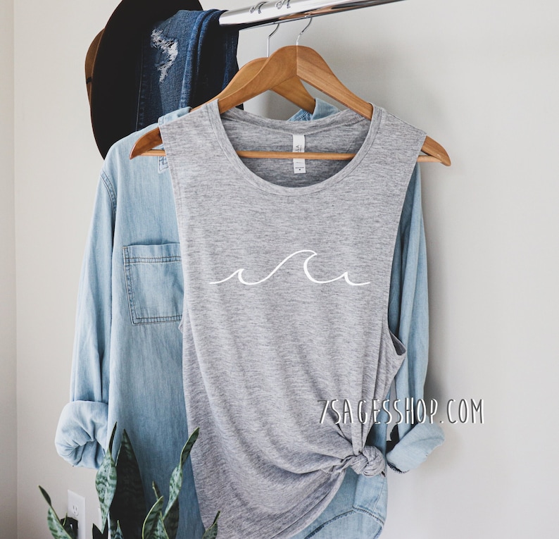 May include: A gray sleeveless muscle tank top with a white wave graphic. The tank top is hanging on a wooden hanger. The text "7sageshop.com" is printed on the bottom right corner of the tank top.