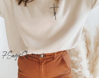 May include: Beige sweatshirt with the word "Faith" in black script. The model is wearing rust-colored pants. The image also shows a necklace and a fluffy, beige decorative element.