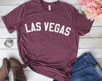 May include: A maroon t-shirt with the text "LAS VEGAS" in white letters. The shirt is laid flat on a white wooden surface with a pair of brown boots and blue jeans.