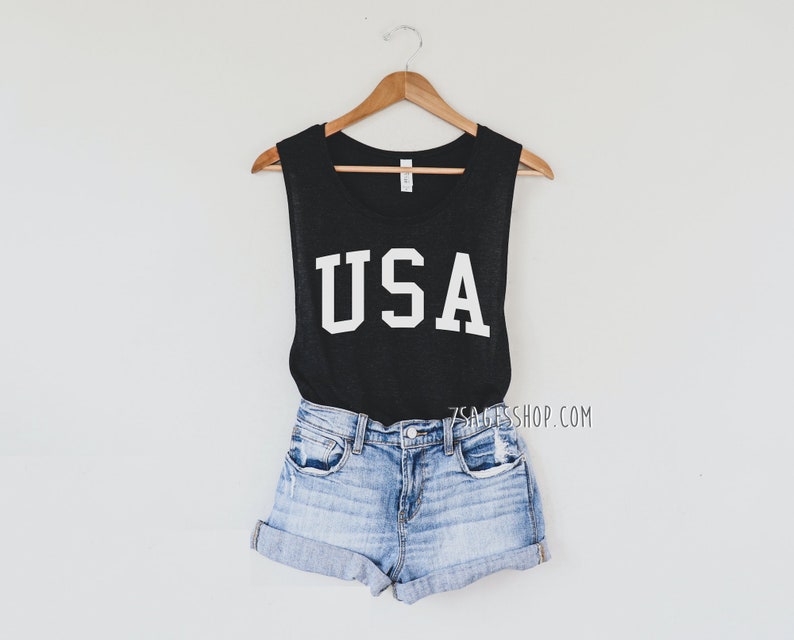 May include: A black sleeveless tank top with the word "USA" in bold white letters. The tank top is paired with light blue denim shorts. The outfit is hanging on a wooden hanger against a white background.