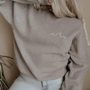 May include: A pale grey sweatshirt featuring a small white wave design. The sweatshirt is paired with light wash jeans. The image is taken in natural light. The website address is visible on the right side of the image.