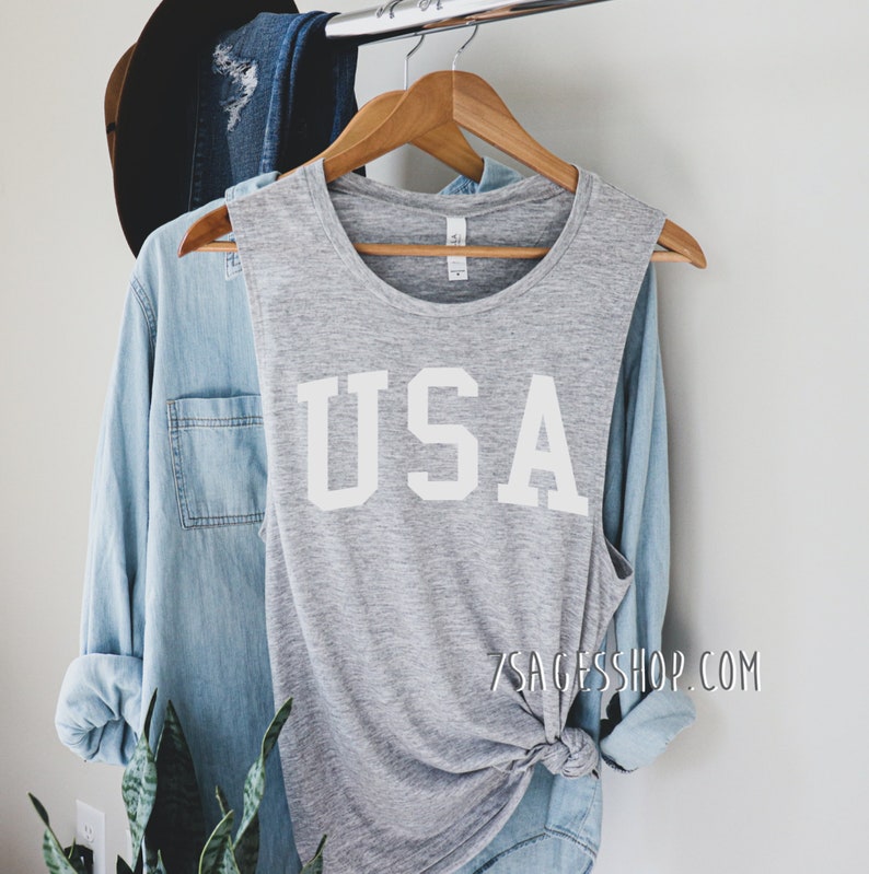 May include: A heather gray tank top with the word "USA" in bold white letters. The tank top is hanging on a wooden hanger, next to a light blue denim shirt and a black hat. The website address 7SAGESSHOP.COM is visible.