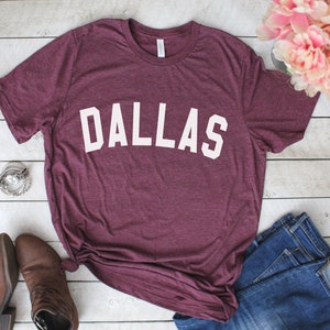 Może przedstawiać: Bordowy t-shirt z nadrukiem "DALLAS" w kolorze białym z przodu.