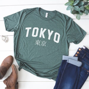 K&ouml;nnte beinhalten: Ein gr&uuml;nes T-Shirt mit dem Wort "TOKYO" in wei&szlig;en Buchstaben und den japanischen Schriftzeichen f&uuml;r "Tokyo" darunter. Das Shirt liegt flach auf einer wei&szlig;en Holzoberfl&auml;che.