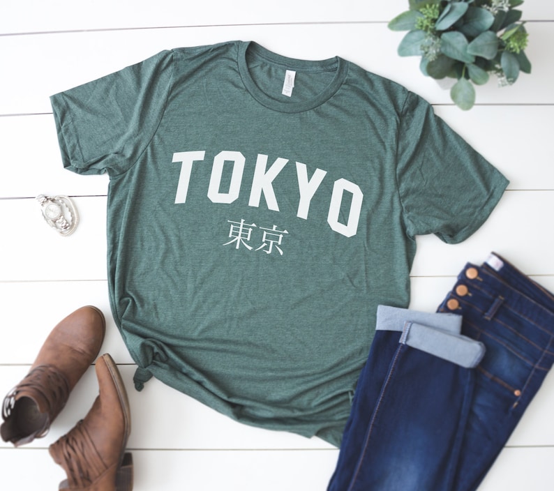 K&ouml;nnte beinhalten: Ein gr&uuml;nes T-Shirt mit dem Wort "TOKYO" in wei&szlig;en Buchstaben und den japanischen Schriftzeichen f&uuml;r "Tokyo" darunter. Das Shirt liegt flach auf einer wei&szlig;en Holzoberfl&auml;che.