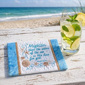 May include: A quilted, rectangular decorative piece with the text "Mightier than the waves of the sea is His love for you" and Psalm 93:4. The piece features embroidered starfish, seashells, and waves, with blue and tan borders. A glass of lemonade with mint and lemon slices sits nearby.