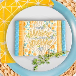 May include: A decorative quilted coaster with the embroidered text "Too Blessed to be Stressed" is placed on a white plate, atop a blue plate. The coaster has a cream background with a floral pattern and is bordered by blue and orange fabric. A yellow napkin and sprig of herbs complete the setting.