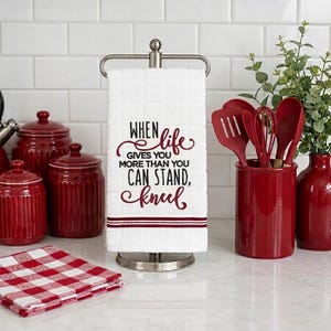 May include: A white waffle-weave kitchen towel hanging on a silver metal stand, featuring the black and red text: When life gives you more than you can stand, kneel. The towel has two thin red horizontal stripes at the bottom edge.