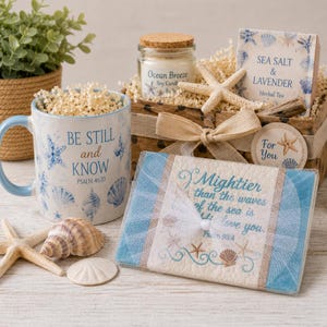 May include: A gift set featuring a ceramic mug with the text BE STILL and KNOW PSALM 46:10, a glass jar candle labeled Ocean Breeze, a box of Sea Salt & Lavender herbal tea, and a rectangular plaque with the text Mightier than the waves of the sea is His love you PSALM 93:4, decorated with starfish and seashells.