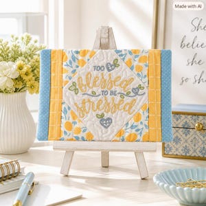 May include: A rectangular quilted fabric wall hanging displayed on a small wooden easel. The center features a diamond-shaped panel with the embroidered text 'TOO BLESSED TO BE STRESSED' in yellow, surrounded by blue floral and yellow lemon patterns.