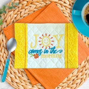 May include: A quilted placemat with the embroidered text "JOY comes in the morning" and a smiling sun design. The placemat is white with yellow borders and is set on an orange napkin and woven placemat. A blue spoon and coffee cup are also present.