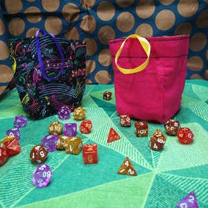 May include: Two fabric drawstring pouches, one black with a colorful musical note pattern and a purple handle, the other solid pink with a yellow handle, surrounded by a collection of multi-sided polyhedral dice in various colors and numbers.
