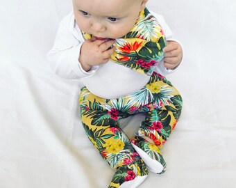 May include: A baby wearing a white long-sleeved onesie, yellow pants, and a matching bib. The pants and bib feature a tropical floral print with red, green, and yellow flowers and leaves. The baby is looking down at the bib.