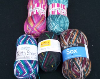 May include: A collection of colorful yarn skeins, including pink, teal, and multicolored options. Some skeins have labels that read "happy", "North Shore Prints", "REGIA", and "Sox". The yarn is arranged on a black surface.