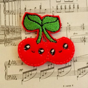 May include: Two red cherries with green stems and leaves, embroidered with smiling faces. The cherries are sewn together and have a white outline.