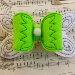 May include: A green felt bow with white felt wings. The wings have a silver glitter design. The bow has a white pom-pom in the center.