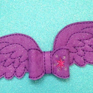 May include: Purple felt wings with a glittery finish and a pink star embroidered on the center. The wings are sewn together in the center to form a bow.