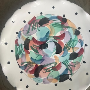 Peut inclure: Une assiette ronde en céramique à bord festonné, dotée d'un rebord blanc orné de pois noirs. Le centre de l'assiette présente un motif abstrait et vibrant composé de coups de pinceau superposés dans des tons de bleu sarcelle, violet, rouge et orange.