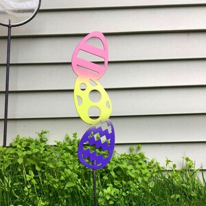 May include: A metal garden stake with three colorful Easter eggs. The eggs are painted in shades of pink, yellow, and purple. The stake is inserted into the ground in a grassy area.