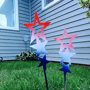 May include: Two metal star yard decorations with a red, white, and blue ombre paint finish. The stars are on metal stakes and are placed in a grassy lawn.