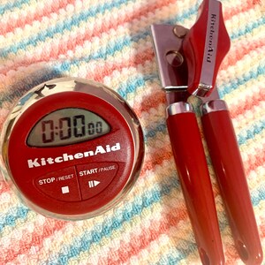 May include: A red KitchenAid digital timer displaying 0:00:00, and a red KitchenAid can opener. The timer has a digital display and control buttons. The can opener has a silver blade and red handles. Both items are on a striped towel.