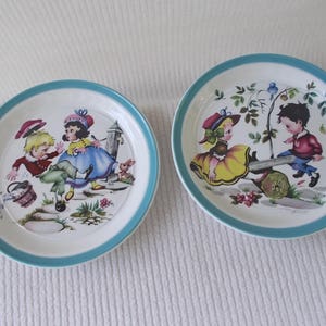 May include: Two vintage children's plates with a blue rim. One plate features a girl in a blue dress and a boy in a red shirt playing with a bucket. The other plate features a girl in a yellow dress and a boy in a red shirt playing on a seesaw.
