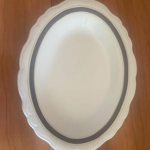 May include: Oval-shaped white serving platter with a scalloped edge. The platter features a dark gray band with a thin gold accent near the center. The design is simple and elegant, suitable for serving food.